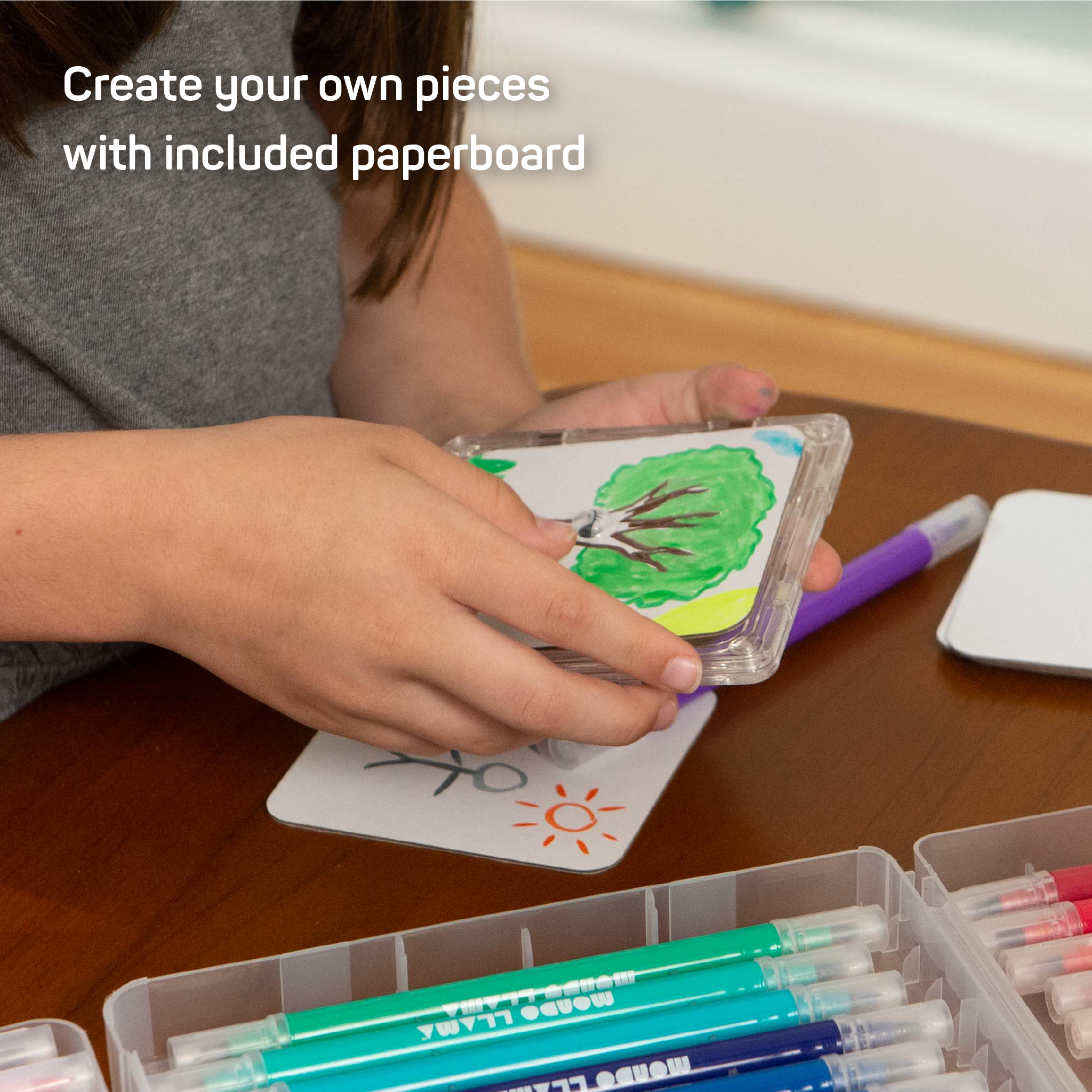 Child placing paperboard panel with drawing of a tree into a square frame. Text which reads "Create your own pieces with included paperboard"